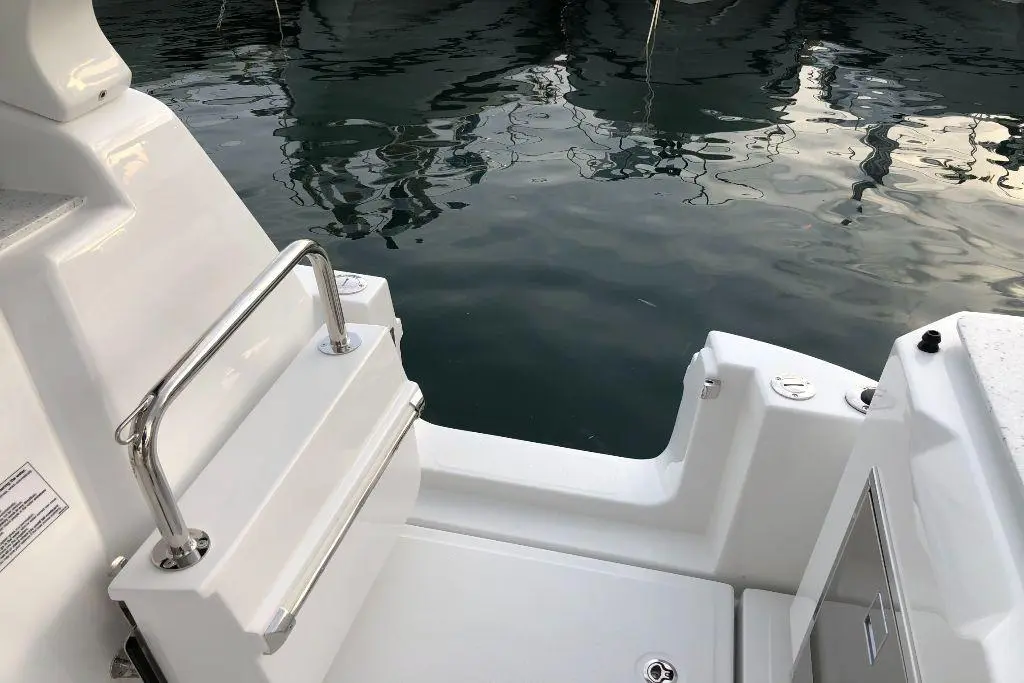 A close-up of the side of a white boat with a stainless steel grab rail and calm water reflecting nearby boats.